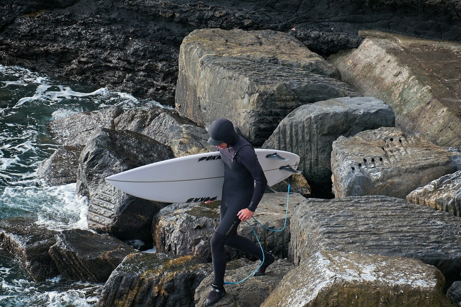 フードとグローブを着けたサーファーが岩場でサーフボードを持っている冬の海岸