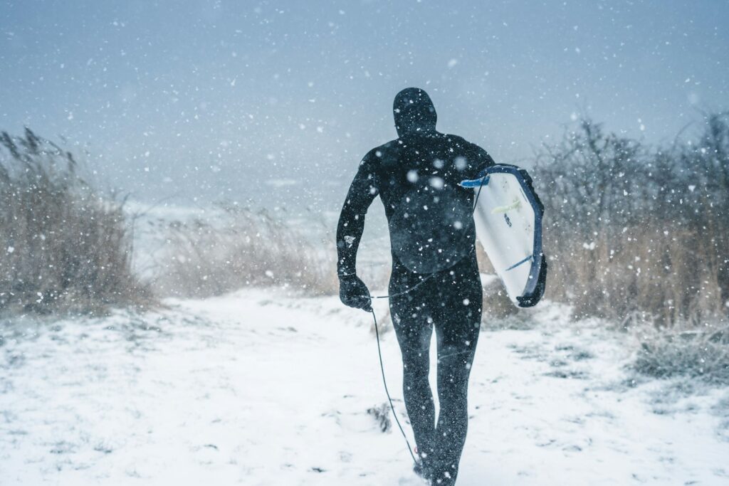 雪の残る海岸をサーフボードを抱えて歩く冬のサーファー｜手荒れ・しもやけ対策