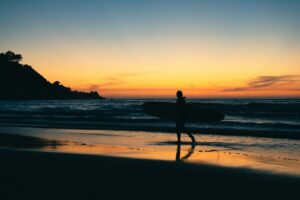 夕暮れの浜をボードを抱えて歩くサーファーのシルエット。ブランク明けの海への一歩